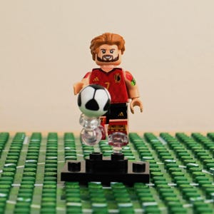 May include: A Lego figurine of a soccer player in a red and black uniform, holding a black and white soccer ball. The figurine has brown hair and a beard. The figure is standing on a black base, with a green field in the background.