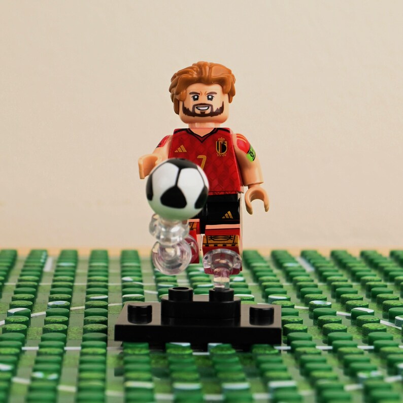 May include: A Lego figurine of a soccer player in a red and black uniform, holding a black and white soccer ball. The figurine has brown hair and a beard. The figure is standing on a black base, with a green field in the background.