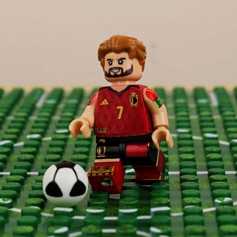May include: A Lego minifigure of a soccer player in a red and black uniform with the number 7. The figure has brown hair and a beard, standing on a green field with a soccer ball.