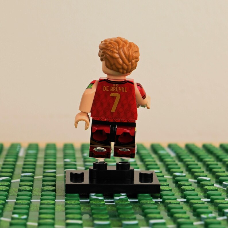 May include: A Lego figurine of a soccer player, viewed from the back. The player wears a red jersey with the number 7 and the name "DE BRUYNE". The figurine has brown hair and is standing on a black base, with a green field in the foreground.