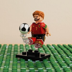 May include: A Lego figurine of a soccer player in a red and black uniform with the number 7. The player is balancing a black and white soccer ball on their foot. The figurine is on a green field.