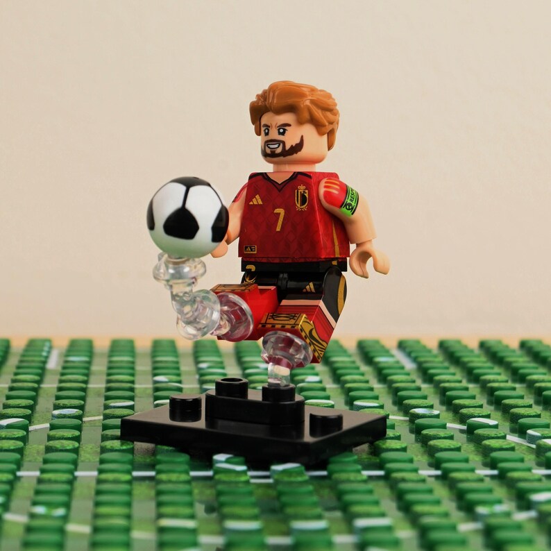 May include: A Lego figurine of a soccer player in a red and black uniform with the number 7. The player is balancing a black and white soccer ball on their foot. The figurine is on a green field.