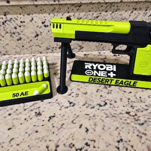 Könnte beinhalten: Eine neongrüne und schwarze Spielzeugpistole auf einem Ständer mit der Aufschrift RYOBI ONE+ DESERT EAGLE, begleitet von einem rechteckigen Tablett mit mehreren neongrünen und weißen Spielzeugpatronen, wobei der Text 50 AE auf dem Tablett steht.
