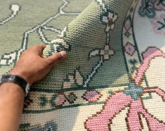May include: A hand-tufted area rug featuring a sage green background with a floral pattern in shades of pink, white, and light blue. The rug has a thick, textured pile and a decorative border with repeating flower motifs and curved ribbon-like shapes.