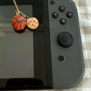 Può includere: Un ciondolo a catena in metallo dorato con una piccola zucca arancione scolpita e un ciondolo a forma di biscotto rotondo, color marrone chiaro e maculato. Il ciondolo è fissato al bordo superiore di una console portatile nera con controller grigio.