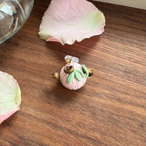Può includere: Un piccolo ciondolo rotondo color rosa chiaro a forma di zucca con scanalature verticali. È decorato con due viticci metallici color bronzo e due foglie verde chiaro sulla parte superiore, con un piccolo anello di sospensione dorato.