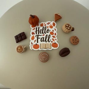 Könnte beinhalten: Ein quadratischer Aufkleber mit dem Text Hello Fall, umgeben von orangefarbenen Kürbissen, Herbstblättern und einem offenen Buch, arrangiert mit Miniatur-Anhängern aus tonartigem Material wie Kürbis, Kuchenstück, Kaffeetassen, Schokolade und Kaffeebohne.