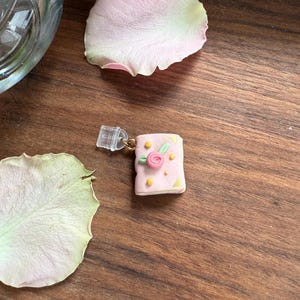 Könnte beinhalten: Ein kleiner, rechteckiger, hellrosa Anhänger in Buchform, verziert mit einer zentralen rosa Rose mit grünen Blättern und kleinen gelben Punkten. Der Anhänger ist mit einem goldfarbenen Metallring an einem transparenten Kunststoffclip befestigt.