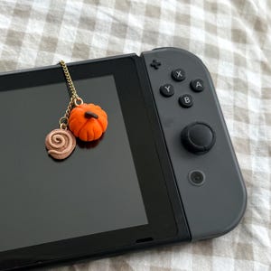Könnte beinhalten: Ein goldfarbener Metallkettenanhänger mit einem kleinen orangefarbenen Kürbis und einem hellbraunen Spiralkeks, der auf dem schwarzen Bildschirm einer tragbaren Spielkonsole mit dunkelgrauem Controller-Teil und A-, B-, X- und Y-Tasten liegt.