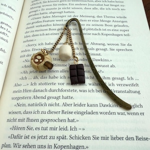Könnte beinhalten: Ein Lesezeichen aus Metall in Antik-Messing-Optik mit drei hängenden Anhängern: einer beigen Tasse mit braunem Herzmotiv, einem weißen zylindrischen Marshmallow und einer dunkelbraunen rechteckigen Schokoladentafel, befestigt an goldfarbenen Ketten.