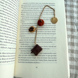 Könnte beinhalten: Ein Lesezeichen aus einer goldfarbenen Metallkette mit drei kleinen Anhängern: einem dunkelbraunen quadratischen Buch, einem beigen Gebäck mit Spiralmuster und einem rostfarbenen Kürbis. Oben an der Kette befindet sich eine flache, runde Scheibe.