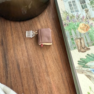 Könnte beinhalten: Ein Miniatur-Buchanhänger in Braun mit einem kleinen roten Lesezeichen-Detail, befestigt an einem transparenten Kunststoff-Bindering. Der Anhänger hat einen glatten, einfarbigen Einband und eine hell abgesetzte Kante für die Buchseiten.