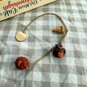 Könnte beinhalten: Ein goldfarbenes Metallkettenarmband mit vier Anhängern: einer schlichten runden Scheibe, einem Stück Kürbiskuchen, einer dunkelbraunen Tasse mit einem kleinen orangefarbenen Kürbis und einem größeren orangefarbenen Kürbis an der Kette.
