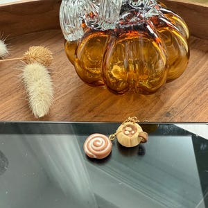 May include: A gold-toned charm bracelet featuring two miniature food-shaped pendants: a round, swirled cinnamon roll and a small, tan-colored mug with a brown handle and a circular design on the front, connected by a delicate gold-colored metal chain.