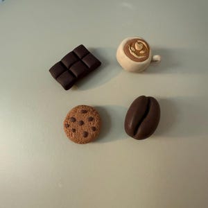 Könnte beinhalten: Ein Set aus vier kleinen Dekoartikeln, bestehend aus einer dunkelbraunen Schokoladentafel, einer hellbeigen Kaffeetasse mit Latte Art, einem runden, hellbraunen Keks mit Schokostückchen und einer einzelnen dunkelbraunen Kaffeebohne auf einer neutralen Fläche.