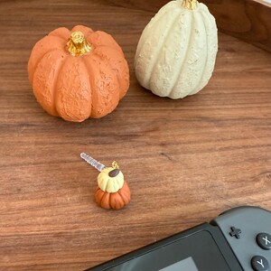 Könnte beinhalten: Ein strukturierter orangefarbener Kürbis mit goldenem Stiel, ein strukturierter cremeweißer Kürbis und ein kleiner Anhänger mit gestapeltem orangefarbenen und cremefarbenen Kürbis mit transparentem Kunststoffstecker, neben einem schwarzen Gamecontroller.