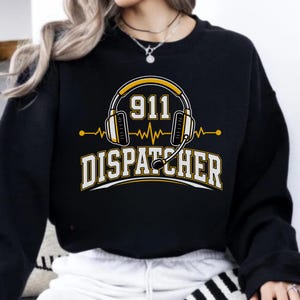 Puede incluir: Una sudadera negra de manga larga con un estampado gráfico de auriculares amarillos y blancos con micrófono. Sobre los auriculares aparece el número 911 y debajo la palabra DISPATCHER en letras blancas en negrita con un borde amarillo.