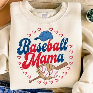 May include: A cream-colored crewneck sweatshirt featuring a graphic print with the text "Baseball Mama" in blue and red script. The design includes a blue baseball cap, a brown baseball glove, two white baseballs, and a ring of red hearts.