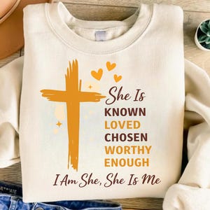 May include: A cream-coloured crewneck sweatshirt featuring a graphic print of a golden-yellow cross, three small hearts, and the text: She Is KNOWN LOVED CHOSEN WORTHY ENOUGH I Am She, She Is Me. The design uses a mix of brown and yellow lettering.