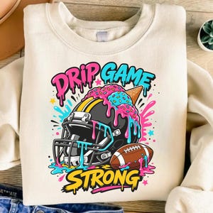 May include: A cream-coloured crewneck sweatshirt featuring a graphic of a black football helmet topped with a melting ice cream cone. The design includes pink and blue drips, a brown football, and the text DRIP GAME STRONG in pink, blue, and yellow.