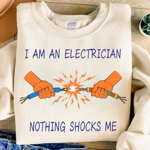 Puede incluir: Una sudadera de cuello redondo color crema con un gráfico de dos manos naranjas sosteniendo cables eléctricos pelados con una chispa naranja brillante en el centro. El texto I AM AN ELECTRICIAN aparece arriba y NOTHING SHOCKS ME abajo en azul.