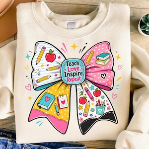 May include: A cream-colored crewneck sweatshirt featuring a large, colorful graphic of a bow. The bow is decorated with school-themed illustrations including pencils, apples, books, and notebooks. The center text reads, Teach, Love, Inspire, Repeat.