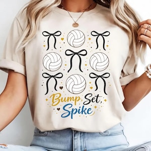 May include: A beige short-sleeve t-shirt featuring a graphic print of four volleyballs and five black bows arranged in a grid. The text "Bump, Set, Spike" is printed in gold and blue script and print lettering below the volleyballs and bows.
