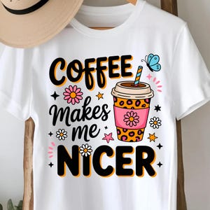 以下が含まれることがあります： ヒョウ柄のコーヒーカップ、ピンクのスリーブ、青いストローのグラフィックプリントが施された白いTシャツ。オレンジ色の縁取りがされた黒い文字で「COFFEE Makes me NICER」と書かれており、花、星、青い蝶のイラストが周囲に配置されています。