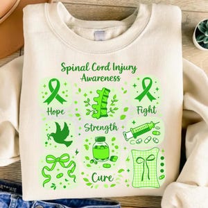 Op de afbeelding: Een crèmekleurig sweatshirt met ronde hals en een groene grafische print met de tekst Spinal Cord Injury Awareness, Hope, Fight, Strength en Cure. Het ontwerp bevat illustraties van linten, een ruggengraat, een duif, een injectiespuit, pillen en een strik.