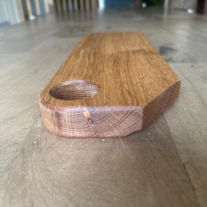 May include: A rectangular wooden serving board with a smooth, light brown finish and a visible natural wood grain pattern. The board features a circular cutout near one end, designed for hanging or handling, with rounded edges and a flat surface.