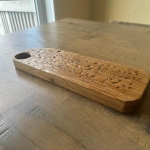 May include: A rectangular wooden serving board with a natural light brown finish and a visible wood grain texture. The board features a circular cutout at one end and a series of small, shallow indentations across the top surface of the wood.