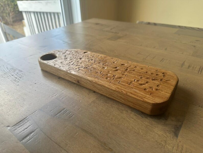 May include: A rectangular wooden serving board with a natural light brown finish and a visible wood grain texture. The board features a circular cutout at one end and a series of small, shallow indentations across the top surface of the wood.