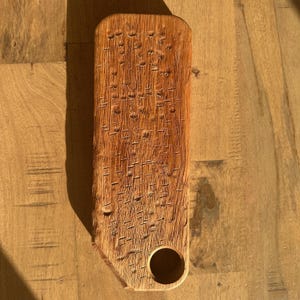 May include: A rectangular wooden board with a natural light brown finish and a prominent wood grain texture. The surface features a series of small, vertical, dash-like indentations across the face and a single circular hole near the bottom corner.