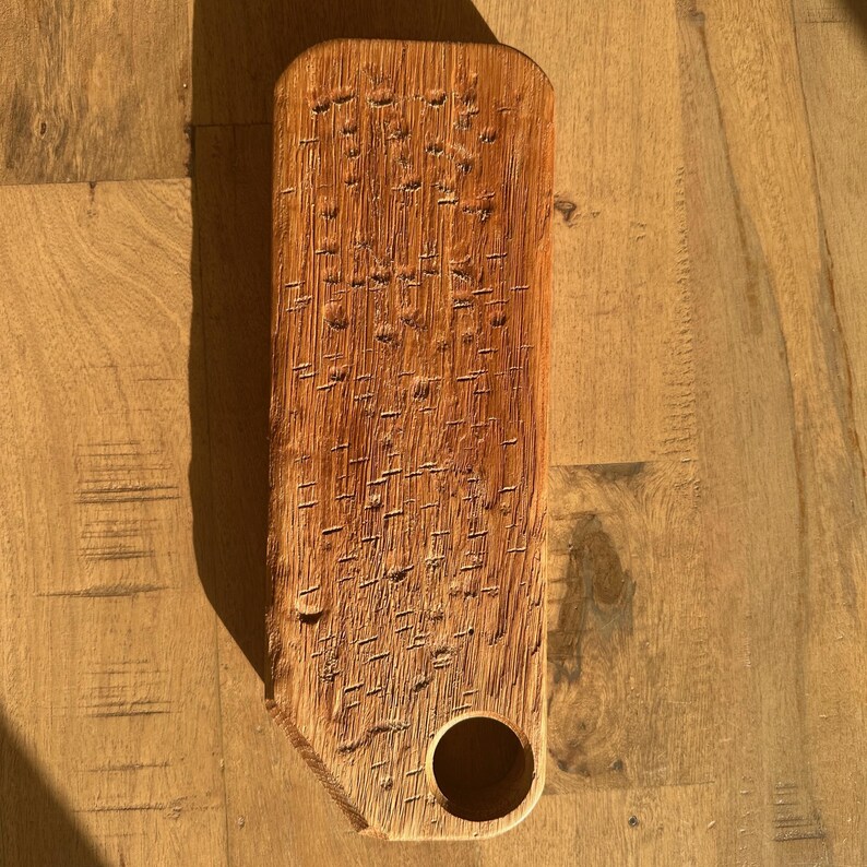 May include: A rectangular wooden board with a natural light brown finish and a prominent wood grain texture. The surface features a series of small, vertical, dash-like indentations across the face and a single circular hole near the bottom corner.