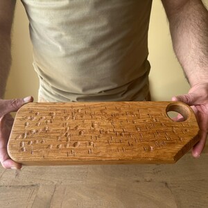 May include: A rectangular wooden serving board with a natural light brown grain and a smooth finish. The surface features a series of small, carved indentations arranged in a scattered pattern. A circular hole is cut into the top right corner.