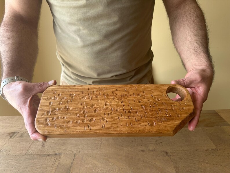 May include: A rectangular wooden serving board with a natural light brown grain and a smooth finish. The surface features a series of small, carved indentations arranged in a scattered pattern. A circular hole is cut into the top right corner.