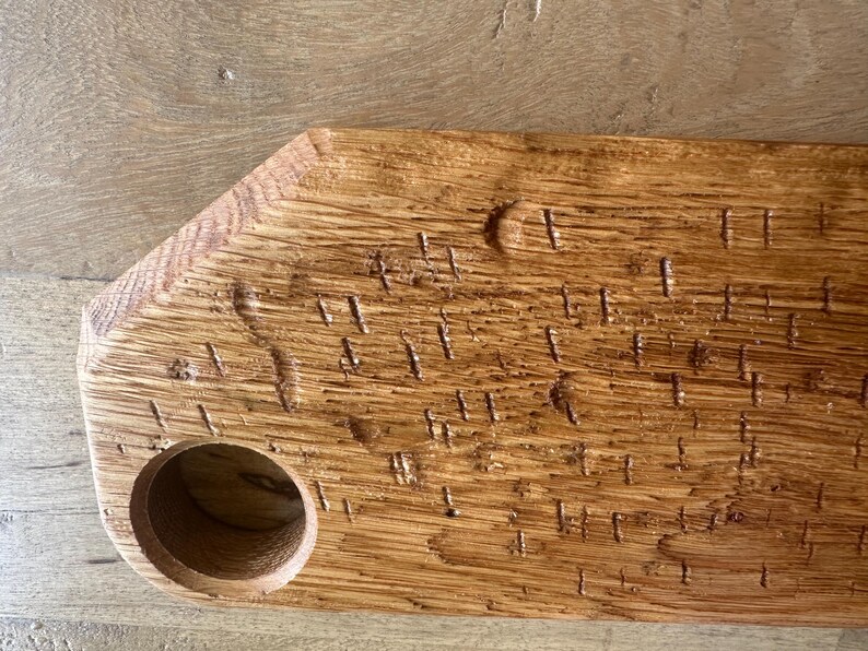 May include: A rectangular wooden serving board with a natural light brown finish and visible wood grain. The board features a circular cutout on the left side and a series of small, vertical, textured indentations across the surface.