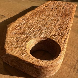 May include: A rectangular wooden serving board with a natural light brown finish and a prominent wood grain texture. The board features a circular cutout near one end, with rough, unfinished edges and a weathered, rustic appearance throughout.