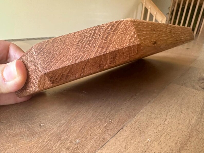 May include: A rectangular piece of reddish-brown oak wood with a visible grain pattern and a beveled edge. The wood block features a smooth, sanded surface texture and a distinct natural wood color, showcasing the dense fibers of the oak material.