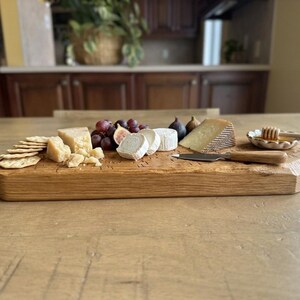 May include: A rectangular wooden serving board featuring a selection of crackers, various cheeses, red grapes, and figs. A small ceramic dish holds honey with a wooden dipper, and a cheese knife with a wooden handle rests on the board surface.