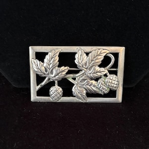 May include: A silver-toned rectangular brooch featuring a detailed botanical design. The brooch showcases a sprig of leaves and pinecones within a rectangular frame. The leaves and pinecones have intricate detailing, and the brooch has a vintage aesthetic.