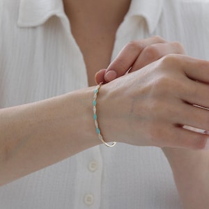 Peut inclure: Un bracelet doré orné de pierres alternées turquoise et transparentes. Le bracelet présente un design délicat, avec de petites pierres turquoises ovales et des pierres transparentes scintillantes. Le bracelet est porté au poignet. Circonférence d'environ 17 cm.