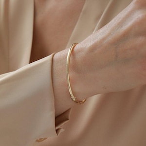 May include: A gold-coloured bangle bracelet is worn on a wrist. The bracelet is a simple, curved design with a clasp. The background is a neutral beige colour.