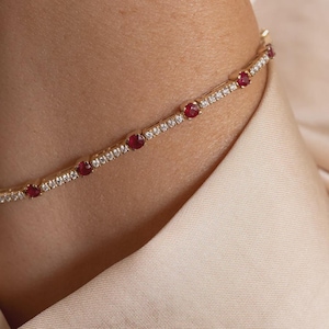 May include: A gold chain bracelet featuring a series of round, deep red rubies interspersed with small, clear, sparkling white diamonds set in a continuous line. The metal band has a polished finish and a secure, delicate link construction.