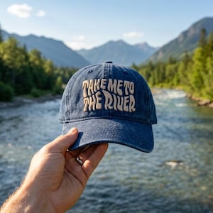 May include: A dark blue denim baseball cap featuring the embroidered text "TAKE ME TO THE RIVER" in a light beige, wavy font across the front panel. The cap has a structured crown, a curved brim, and a classic six-panel design with stitching.