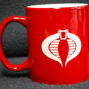 May include: A red ceramic coffee mug with a white Cobra logo. The logo is a stylized cobra with a crown on its head.