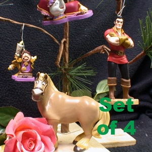 May include: Set of four Disney Beauty and the Beast Christmas decorations. The decorations feature Belle, Beast, Gaston, and the horse, Philippe. The decorations are hanging from a small artificial pine tree.
