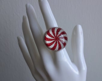 May include: A red and white candy cane swirl ring with glitter embedded in the resin. The ring is on a white mannequin hand.