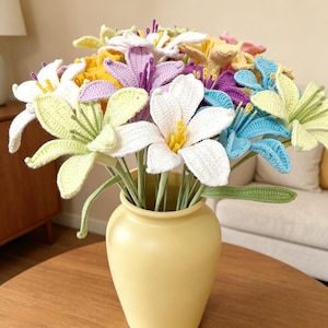 Könnte beinhalten: Ein Strauß aus gehäkelten Lilienblüten in verschiedenen Farben wie Weiß, Hellgelb, Lavendel und Blau, jeweils mit einem zentralen Staubblatt, arrangiert in einer glatten, matten gelben Keramikvase mit rundem Körper und engem Hals.
