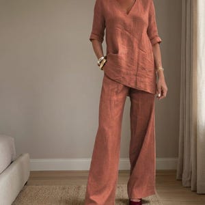 May include: A rust-colored linen outfit consisting of a V-neck top with a pocket and wide-leg pants. The top has short sleeves. The outfit is paired with gold bracelets and dark red shoes. The outfit is made of a lightweight fabric.